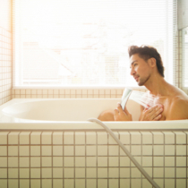 Asian man showering in modern Japanese bathtub, back lit by window.
